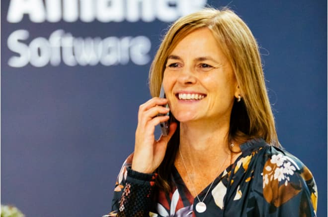 Kristie, an Alliance project manager, talking on the phone, pictured in front of a wall with the Alliance Software logo